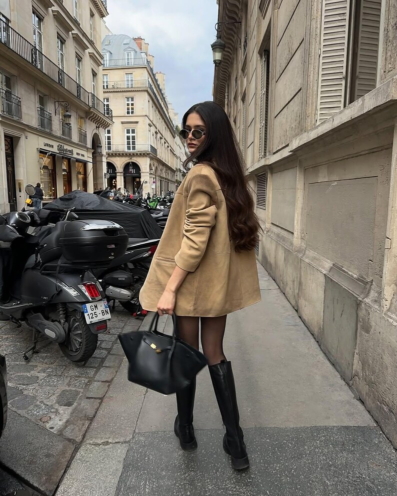 Pu&ograve; includere: Una donna con un blazer beige, collant neri e stivali neri alti fino al ginocchio cammina per una strada cittadina. Porta una borsa a mano in pelle nera.