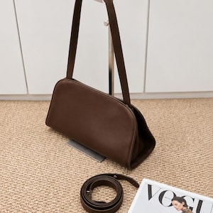 May include: A brown leather shoulder bag with a matching strap, displayed on a metal stand. A rolled-up belt and a magazine are next to the bag. The text "KarachiLeatherCo" is visible at the bottom.