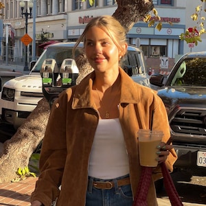 May include: A woman wearing a brown suede jacket, a white tank top, blue jeans, and a brown leather belt. She is holding a brown paper cup with a straw and a dark red woven bag. She is standing in front of a building with a sign that says "Jewelers".