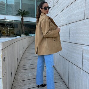 May include: A tan suede jacket with a collared neckline and long sleeves paired with light blue denim jeans featuring distressed knee detailing. The jacket has a button-front closure and a straight hemline, while the jeans have a straight-leg cut.
