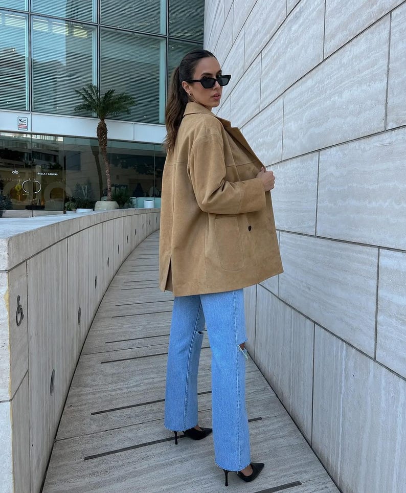 May include: A tan suede jacket with a collared neckline and long sleeves paired with light blue denim jeans featuring distressed knee detailing. The jacket has a button-front closure and a straight hemline, while the jeans have a straight-leg cut.