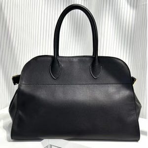 Puede incluir: Un bolso de mano de cuero negro con una silueta estructurada de parte superior redondeada y dos asas verticales fijas. El bolso presenta un acabado liso y mate, una costura horizontal en la sección superior y herrajes metálicos dorados laterales.