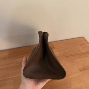 Op de afbeelding: Een bruine leren tas met een driehoekig silhouet en een opening aan de bovenzijde. De tas is gemaakt van glad, effen materiaal met zichtbare stiksels langs de randen en heeft een gestructureerde vorm die naar boven toe smaller toeloopt.