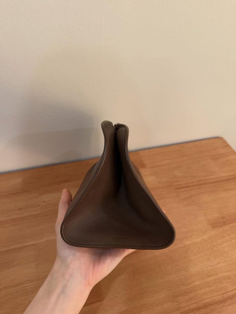 Op de afbeelding: Een bruine leren tas met een driehoekig silhouet en een opening aan de bovenzijde. De tas is gemaakt van glad, effen materiaal met zichtbare stiksels langs de randen en heeft een gestructureerde vorm die naar boven toe smaller toeloopt.