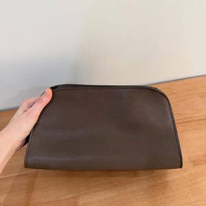 Op de afbeelding: Een donkerbruine, semi-gestructureerde tas met een gladde, matte afwerking. De tas heeft een gebogen bovenrand, een ritssluiting langs de bovennaad en zichtbare donkere stiksels langs de randen, wat zorgt voor een eenvoudig en functioneel ontwerp.