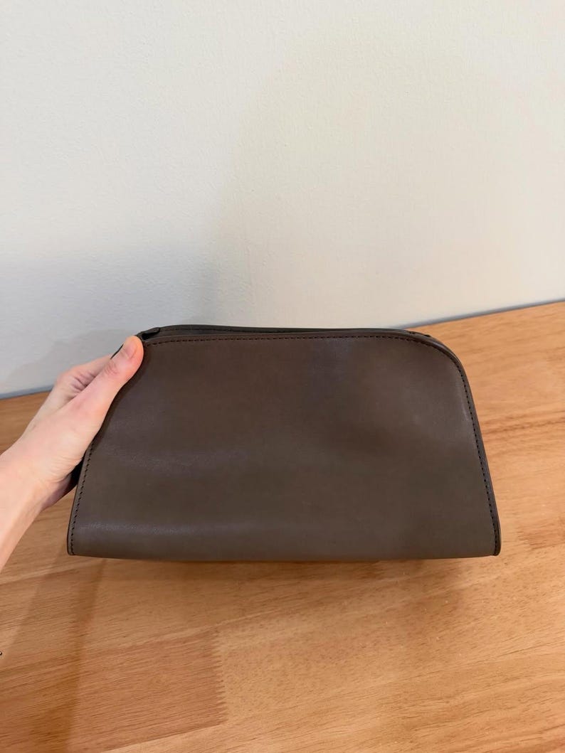 Op de afbeelding: Een donkerbruine, semi-gestructureerde tas met een gladde, matte afwerking. De tas heeft een gebogen bovenrand, een ritssluiting langs de bovennaad en zichtbare donkere stiksels langs de randen, wat zorgt voor een eenvoudig en functioneel ontwerp.