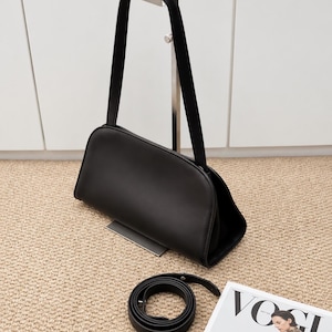 Op de afbeelding: Een zwarte leren handtas met een glad, gebogen silhouet aan de bovenkant en een enkele schouderband. Een bijpassende zwarte leren riem ligt opgerold op het oppervlak eronder. De tekst KarachiLeatherCo staat onderaan in het beeld.