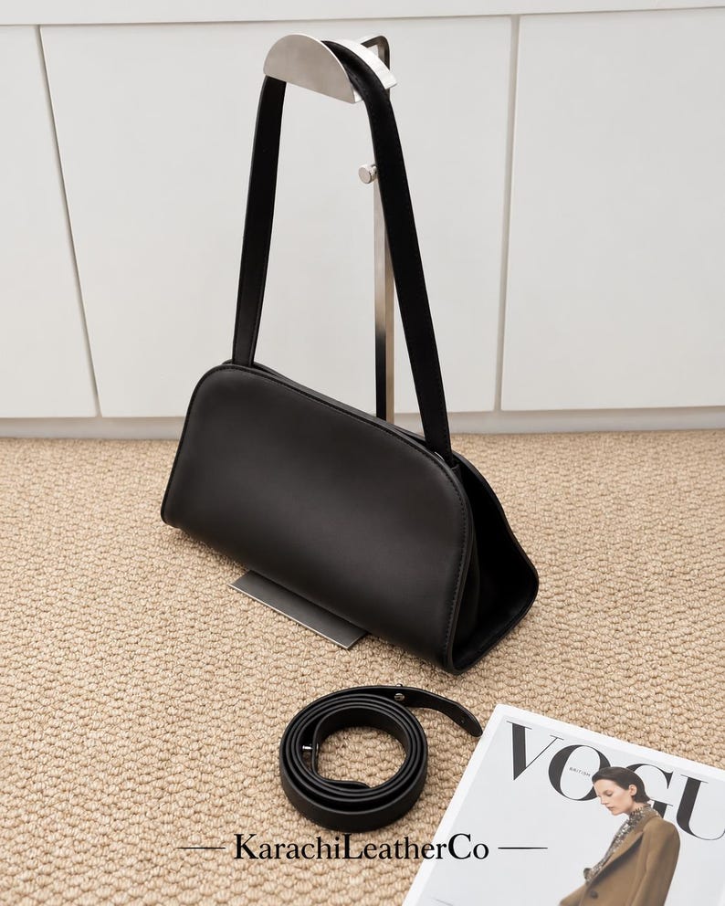 Op de afbeelding: Een zwarte leren handtas met een glad, gebogen silhouet aan de bovenkant en een enkele schouderband. Een bijpassende zwarte leren riem ligt opgerold op het oppervlak eronder. De tekst KarachiLeatherCo staat onderaan in het beeld.