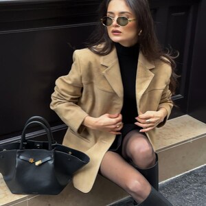Pu&ograve; includere: Una donna indossa un blazer in pelle scamosciata color beige, un maglione nero a collo alto, collant neri e stivali neri. Tiene una borsa tote in pelle nera con una fibbia dorata.