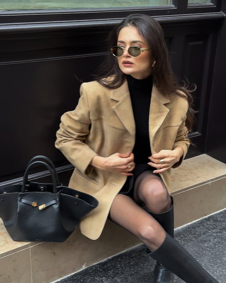 Pu&ograve; includere: Una donna indossa un blazer in pelle scamosciata color beige, un maglione nero a collo alto, collant neri e stivali neri. Tiene una borsa tote in pelle nera con una fibbia dorata.