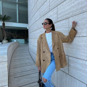 May include: A tan suede double-breasted blazer with black buttons, worn over a plain white t-shirt and light blue distressed denim jeans. The outfit is paired with dark rectangular sunglasses and a black patterned shoulder bag with a strap.