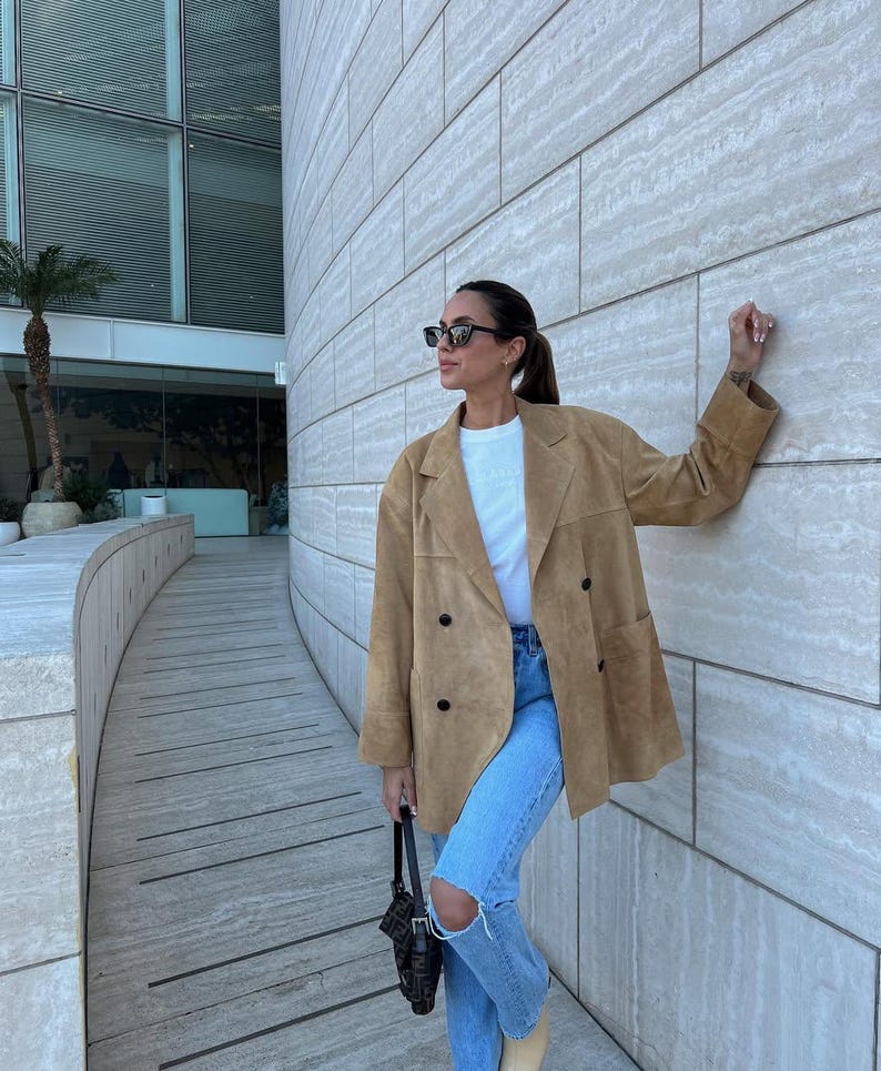 May include: A tan suede double-breasted blazer with black buttons, worn over a plain white t-shirt and light blue distressed denim jeans. The outfit is paired with dark rectangular sunglasses and a black patterned shoulder bag with a strap.