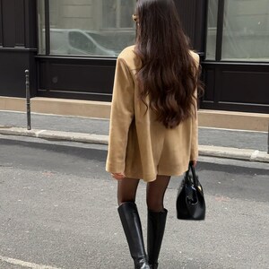 Pu&ograve; includere: Una donna con una giacca di camoscio marrone e stivali neri alti fino al ginocchio cammina per una strada cittadina. Porta una borsa di pelle nera.