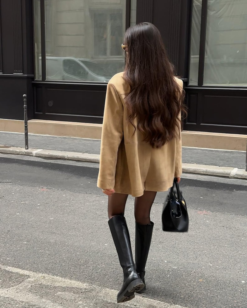 Pu&ograve; includere: Una donna con una giacca di camoscio marrone e stivali neri alti fino al ginocchio cammina per una strada cittadina. Porta una borsa di pelle nera.