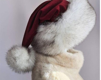 Puede incluir: Sombrero de Papá Noel de terciopelo burdeos con ribete y borla de piel sintética blanca. El sombrero lo lleva puesto una persona que lleva un abrigo de piel blanca.