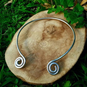 May include: Silver choker necklace with two spiral ends. The necklace is on a wooden surface.