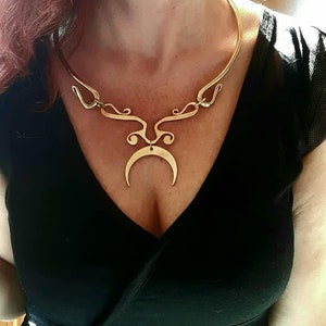 Puede incluir: Un collar gargantilla dorado con un colgante de luna creciente. El collar está hecho de metal grueso y tiene un diseño decorativo.