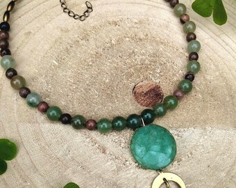 May include: A beaded necklace featuring green, brown, and gold beads. A large, round, green pendant hangs from the necklace, along with a gold-colored crescent moon and teardrop charm. The necklace is displayed on a wooden surface.