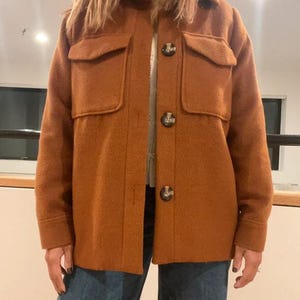 May include: A rust-colored jacket with two chest pockets and three large buttons down the front. The jacket is made of a textured fabric and is worn over a lighter-colored top and blue jeans. The jacket has a collared neckline.