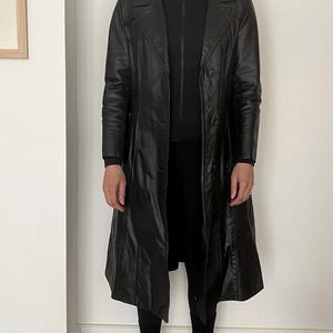 May include: A long, black leather coat with a notched collar and two-button closure. The coat has a tailored fit and falls to the mid-calf. The coat is worn over a black outfit, creating a sleek, monochromatic look.