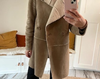 May include: A tan coat with a cream-colored shearling lining and collar. The coat has a double-breasted design with two buttons and a long length. The sleeves are long, and the coat is worn open. The coat is made of a soft, suede-like material.