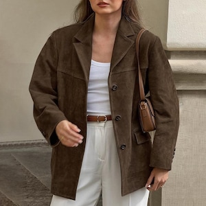 May include: A brown suede blazer with a notched collar and button fastening. The blazer is worn with a white vest top, white trousers, and a brown leather belt. A small brown handbag hangs from the shoulder.