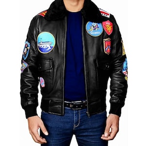 Può includere: Giacca bomber in pelle nera con diverse toppe ricamate. La giacca presenta un colletto in pelliccia nera, due tasche frontali e polsini e orlo a coste neri. Indossata sopra una maglia blu e jeans blu.