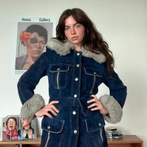 May include: A dark blue suede jacket with a faux fur collar and cuffs. The jacket has a belted waist, four front pockets, and silver button closures. The jacket is worn by a person in front of a wall with a poster.
