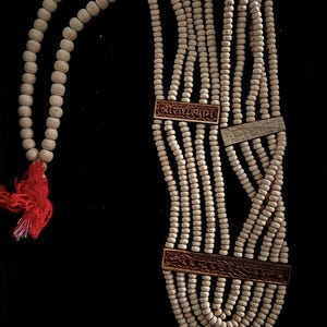 May include: A multi-strand necklace with light-colored wooden beads and decorative rectangular wooden accents. The necklace has a red tassel and features engraved text in a script font. The background is black.