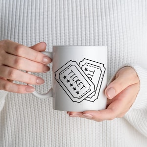 May include: White ceramic mug with a black and white design of two vintage-style tickets. The word "TICKET" is printed on one ticket, with stars. The mug is held by a person wearing a white jumper.