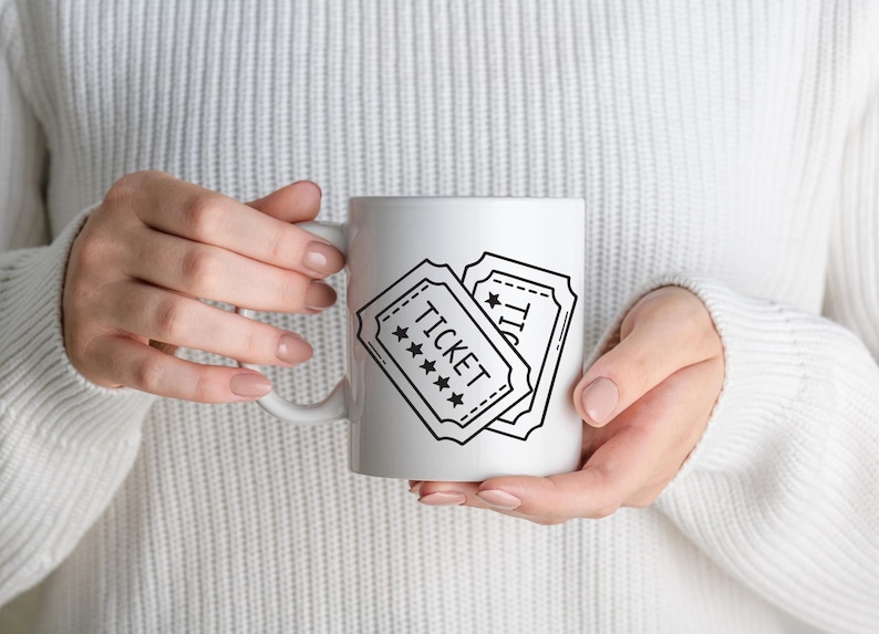 May include: White ceramic mug with a black and white design of two vintage-style tickets. The word "TICKET" is printed on one ticket, with stars. The mug is held by a person wearing a white jumper.