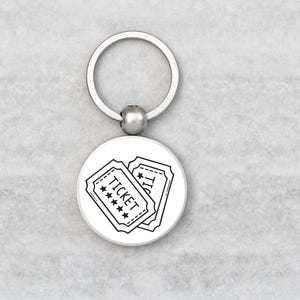 May include: A silver keyring with a circular charm displaying two black and white ticket illustrations. The tickets have the word "TICKET" and star details. The keyring is on a light grey surface.