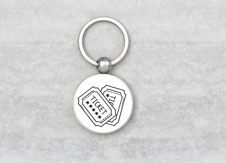 May include: A silver keyring with a circular charm displaying two black and white ticket illustrations. The tickets have the word "TICKET" and star details. The keyring is on a light grey surface.