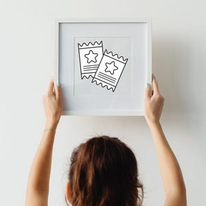 May include: A white framed print featuring two black and white tickets with star designs. The tickets have a serrated edge and horizontal lines. The print is held by a person with light skin and a gold bracelet, against a white wall.