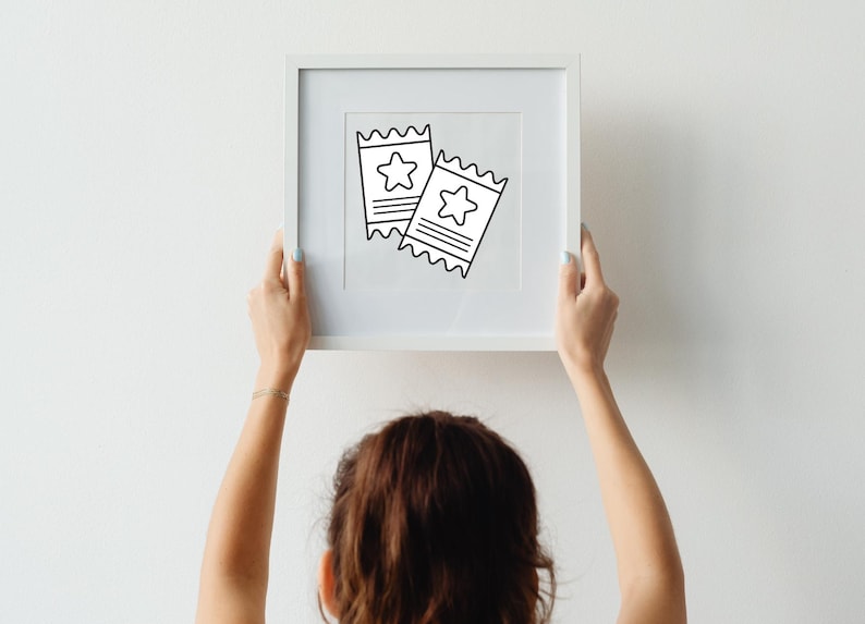 May include: A white framed print featuring two black and white tickets with star designs. The tickets have a serrated edge and horizontal lines. The print is held by a person with light skin and a gold bracelet, against a white wall.