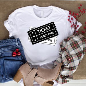 May include: White t-shirt featuring a black ticket graphic with the text "TICKET ADMIT ONE". The image includes blue jeans, tan boots, and a plaid scarf. Red baubles and berries add a festive touch.