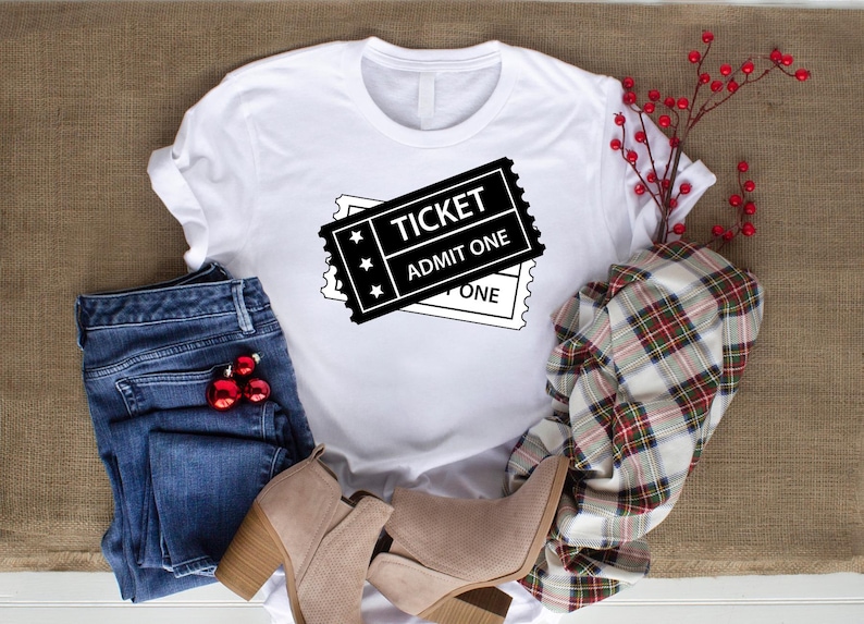 May include: White t-shirt featuring a black ticket graphic with the text "TICKET ADMIT ONE". The image includes blue jeans, tan boots, and a plaid scarf. Red baubles and berries add a festive touch.
