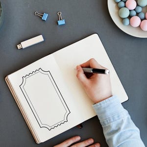 May include: An open notebook with a hand sketching a decorative frame with a pen. A small succulent plant, a USB drive, and a plate of pastel sweets are also on the grey surface. Two blue binder clips are visible.