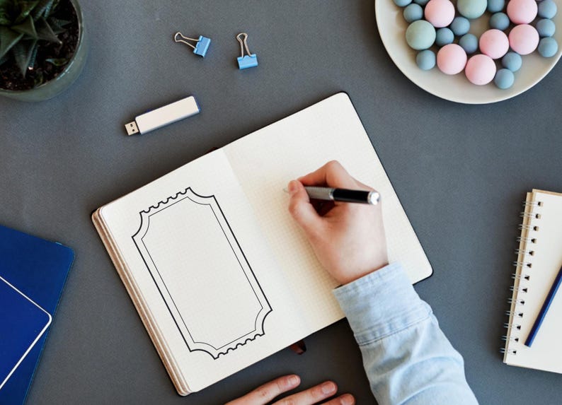 May include: An open notebook with a hand sketching a decorative frame with a pen. A small succulent plant, a USB drive, and a plate of pastel sweets are also on the grey surface. Two blue binder clips are visible.