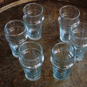 May include: Seven clear glass drinking glasses with a textured surface sit on a gold tray. The glasses are all different sizes and shapes.