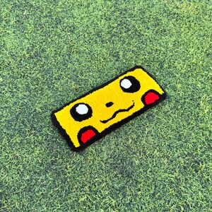 May include: A rectangular yellow rug with a Pikachu cartoon face, black outlines, white eyes, and red cheek accents. The rug is on a green grassy surface.