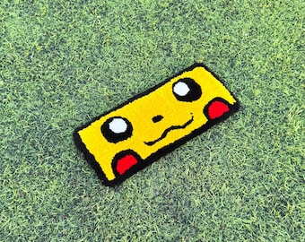 Può includere: Un tappeto rettangolare giallo con la faccia di Pikachu, contorni neri, occhi bianchi e guance rosse. Il tappeto è posto su una superficie erbosa verde.
