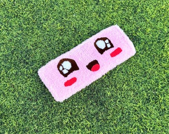 May include: A rectangular pink rug with a cartoon face design. The rug features brown eyes, a black mouth, and red cheeks. The rug is placed on green grass.