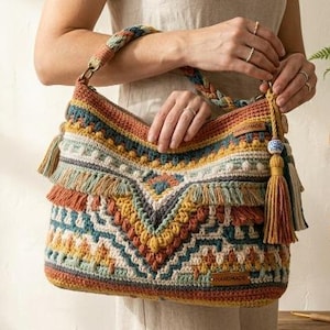 May include: A crochet handbag featuring a multi-colored geometric pattern in shades of rust, teal, mustard yellow, and cream. The bag includes a braided handle, decorative fringe along the front, and a long tassel accent with a blue and white bead.