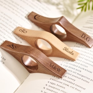 May include: Three wooden thumb page holders in varying shades of brown and tan, resting on an open book. Each holder features a unique design: a mountain, an infinity heart, and a crescent moon. Some are engraved with initials or a name.