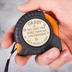 May include: A black and orange tape measure with a wooden centre. The wooden centre is engraved with the words "DADDY WE LOVE YOU MORE THAN WE CAN MEASURE FROM SOPHIA & LIBBY" and star accents.