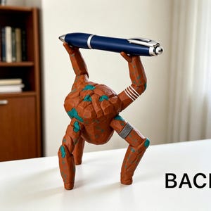 May include: A brown, rock-textured creature figurine with teal accents standing on three legs. The figure is balancing a dark blue ballpoint pen with a silver clip and tip horizontally above its head. The word BACK is printed in black.
