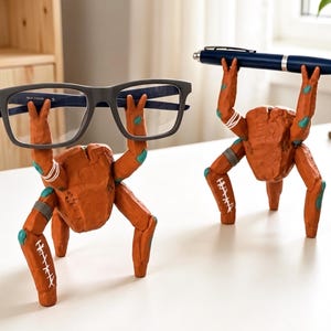 May include: Two orange, textured, multi-legged creature figurines acting as desk organizers. One figure holds a pair of dark gray eyeglasses, while the other holds a dark blue ballpoint pen. Both figures feature white stitched patterns and teal accents.