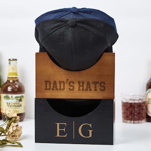 Könnte beinhalten: Ein hölzerner Hut-Organizer mit dem Text "DAD'S HATS" und den Initialen "E|G". Der Organizer hält eine schwarze und marineblaue Baseballkappe. Bierflaschen und ein Glas Flüssigkeit befinden sich im Hintergrund.