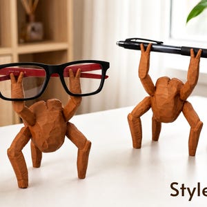 May include: Two brown, carved wooden figures shaped like multi-legged creatures. One figure holds a pair of black-framed eyeglasses with red inner temples, while the other holds a black ballpoint pen. The text Style B is visible at the bottom.